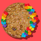 Specialty cookie cake decorated with rainbow frosting, colorful sprinkles, and a golden-brown chocolate chip base on a red background.