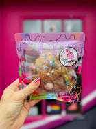 Specialty lactation cookie with chocolate, marshmallows, and colorful sprinkles, in Rebel Daughter Cookies packaging, held against a vibrant pink background.