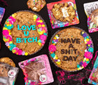 Custom cookie cake display featuring two decorated chocolate chip cookie cakes with bold messages, colorful frosting, and sprinkles, surrounded by packaged cookies.