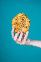 A woman's hand holding a gourmet chocolate chunk walnut Rebel Daughter cookie against a teal background