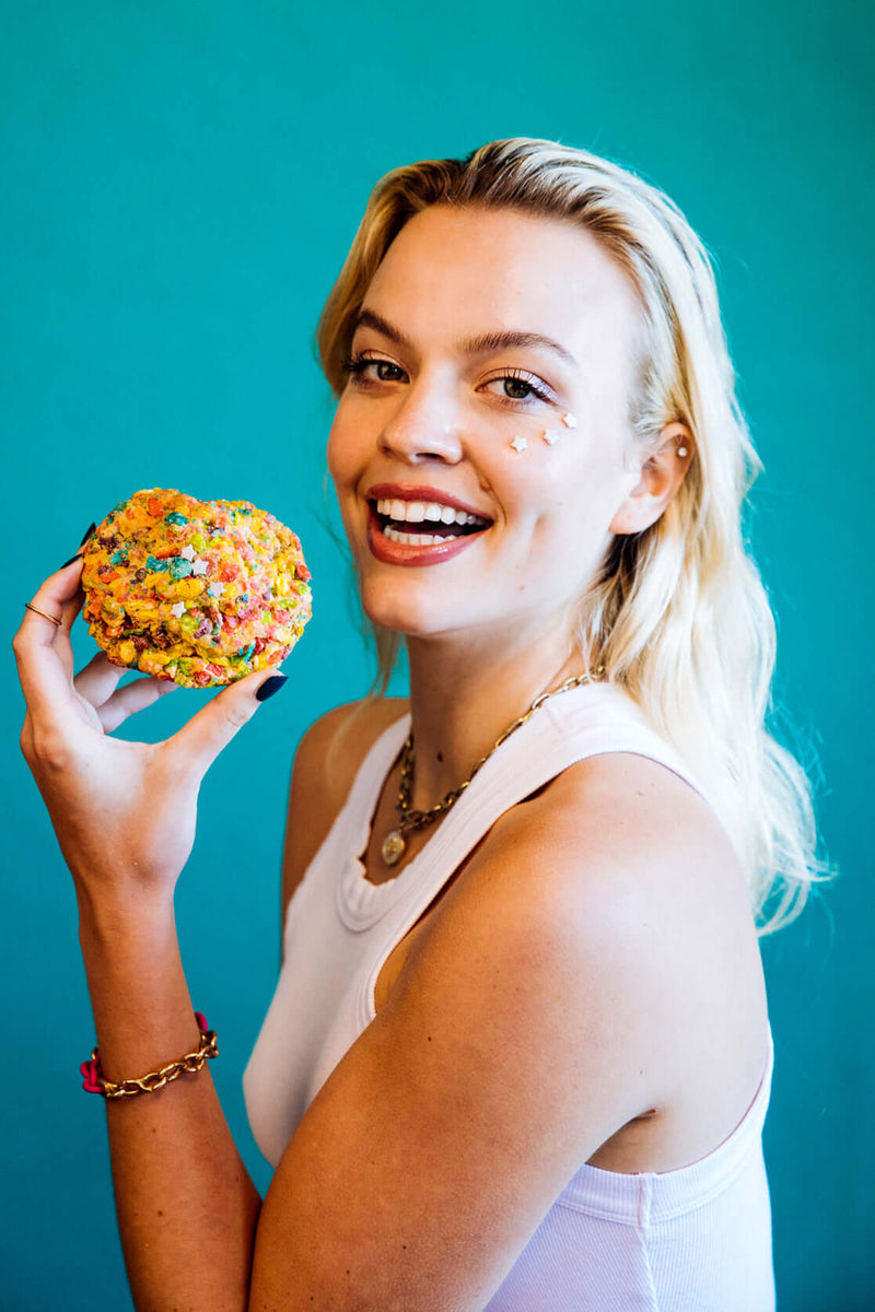 A blonde model holding a colorful, gourmet Rebel Daughter White Unicorn cookie covered in Fruity Pebbles against a teal background