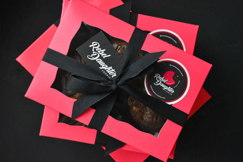 A stack of 3 pink Rebel Daughter Cookies delivery boxes against a black background