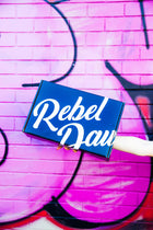 A hand holding a black and white Rebel Daughter Cookies shipping box against a pink mural
