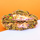 A colorful, gourmet Rebel Daughter White Unicorn cookie covered in Fruity Pebbles cut in half and stacked against an orange and white background
