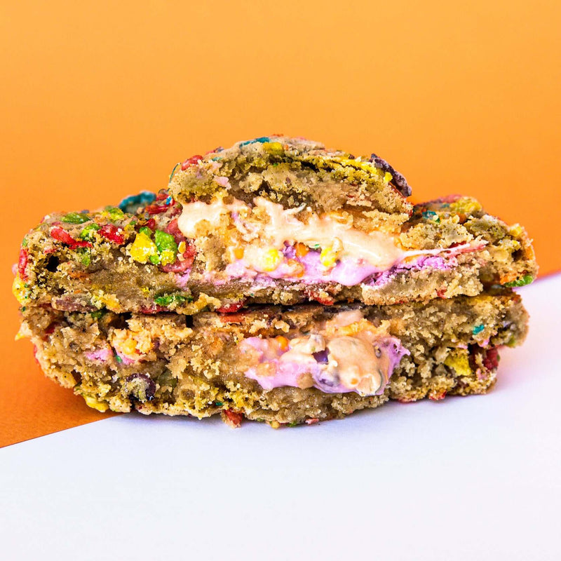 A colorful, gourmet Rebel Daughter White Unicorn cookie covered in Fruity Pebbles cut in half and stacked against an orange and white background