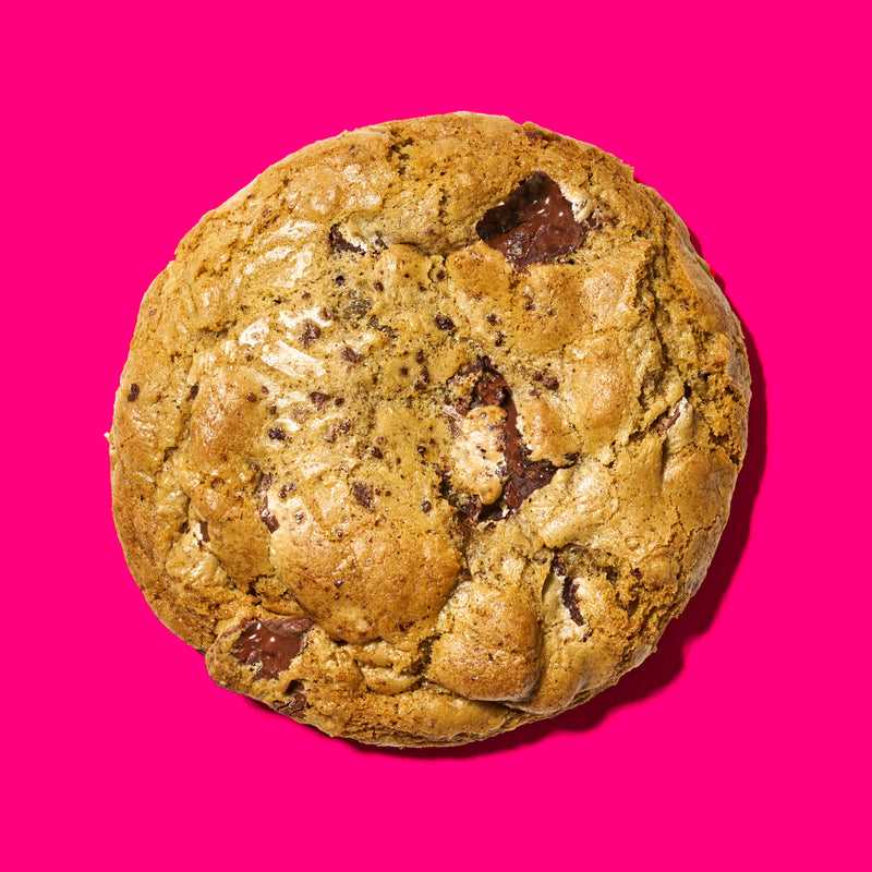 Gourmet chocolate cookie with a golden-brown exterior and melted chocolate chunks on a vibrant pink background.