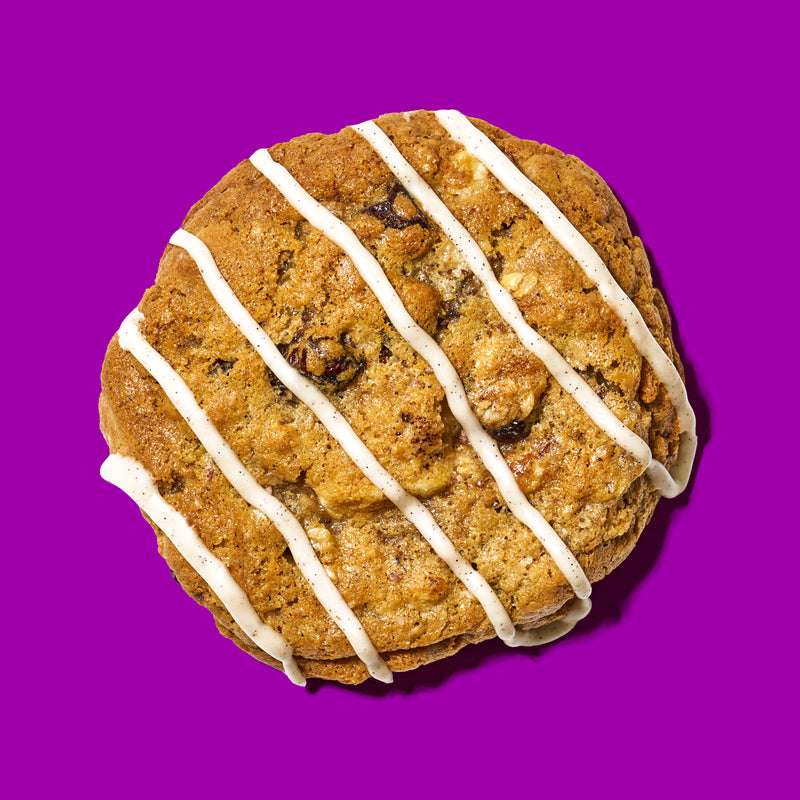 Oatmeal and raisins specialty cookie with a golden-brown texture, drizzled with vanilla icing on a bold purple background.