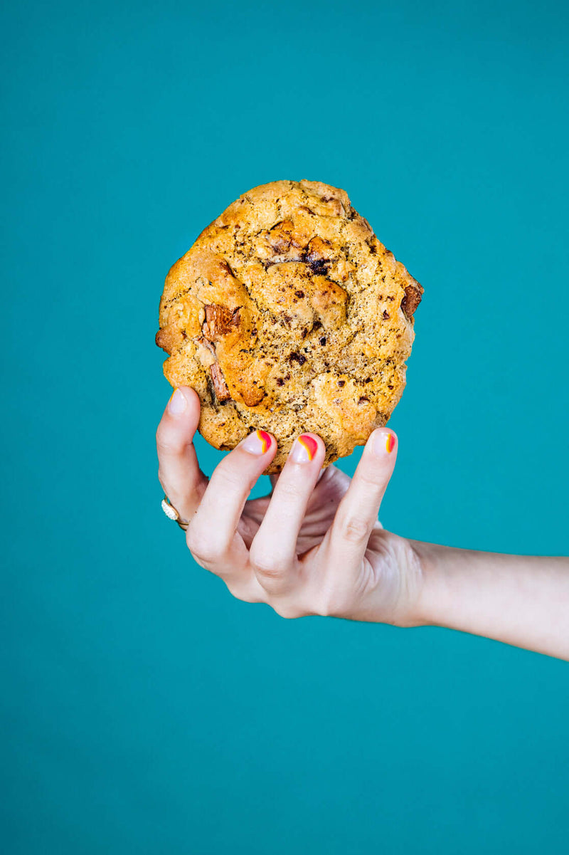 A woman's hand holding a gourmet chocolate chunk walnut Rebel Daughter cookie against a teal background