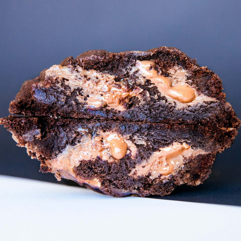 A gooey Rebel Daughter gourmet chocolate chunk caramel brownie cookie cut in half and stacked against a navy blue and white background
