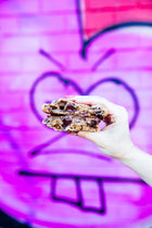 A woman's hand holding a gourmet chocolate chunk walnut Rebel Daughter cookie cut in half and stacked against a purple graffiti mural