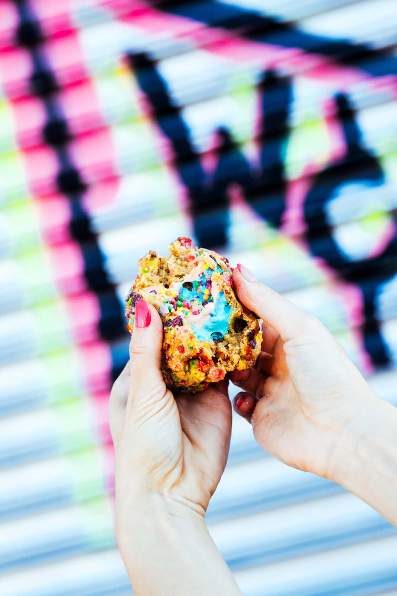 A colorful, gourmet Rebel Daughter White Unicorn cookie being broken in half showing a colorful candy center, against a graffiti mural