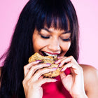 A woman biting into a Rebel Daughter chocolate chunk cookie against a pink background