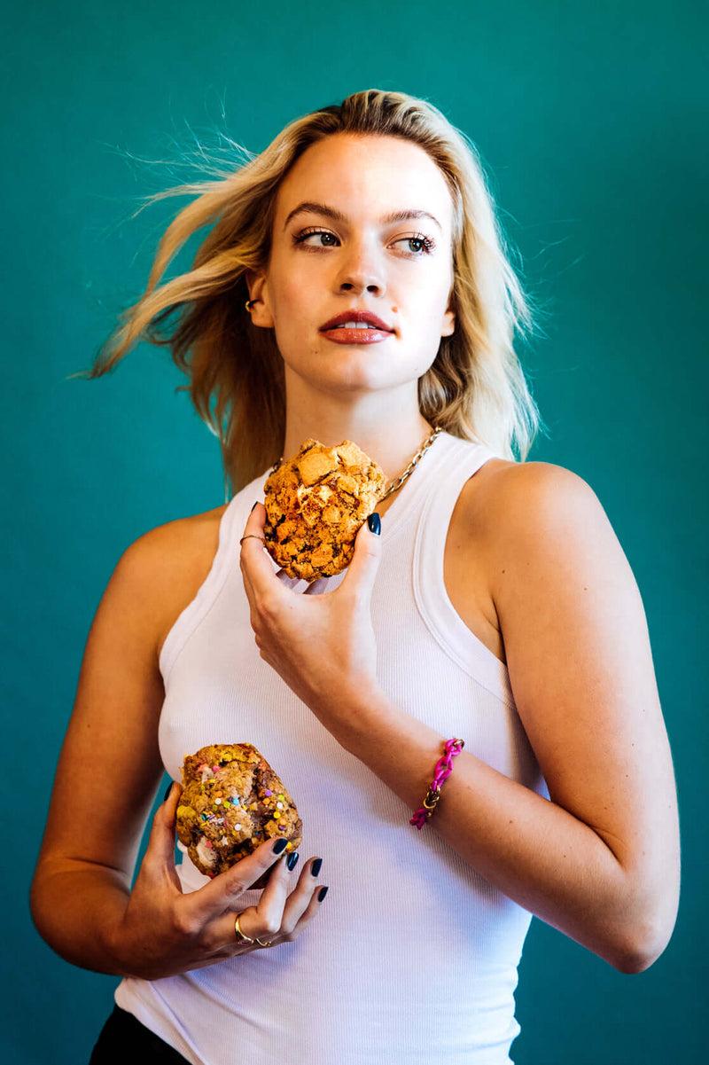 A blonde model in a white tank top holding Rebel Daughter Cookies against a teal background